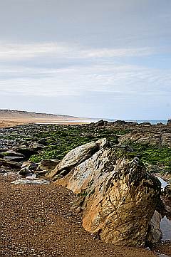 Steilküste mit Felsbrocken im groben Sand unter blauem Himmel